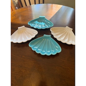 May include: A set of four ceramic dishes shaped like scallop shells, featuring two in a glossy turquoise blue finish and two in a glossy white finish. Each dish has a fan-like design with raised ridges radiating from the base to the edge.