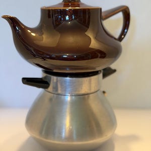 May include: A two-part coffee maker featuring a glossy dark brown ceramic teapot with a matching lid and handle, resting securely on a brushed silver-tone metal base with black plastic side handles and a rounded, bulbous lower body shape.