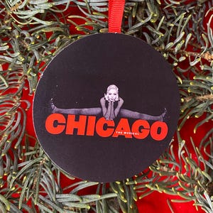 May include: A circular black ornament featuring a graphic of a performer doing a split with the red text CHICAGO THE MUSICAL printed below. The ornament is suspended by a thin red ribbon loop at the top, centered against a dark background.
