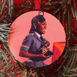 May include: A circular ornament featuring a printed image of a person with braided hair wearing a grey blazer with red, white, and blue sleeve stripes, smiling while holding a gold gramophone trophy against a warm, reddish-orange background.