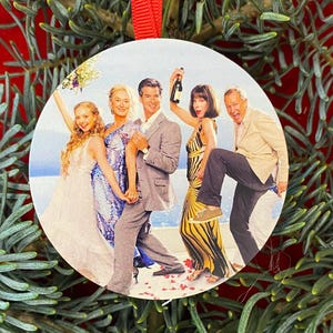 May include: A round, flat Christmas ornament featuring a printed image of five people from the movie Mamma Mia. The group includes a person in a white dress, a person in a blue patterned dress, a person in a grey suit, a person in a yellow and black striped dress holding a bottle, and a person in a tan blazer and dark trousers.