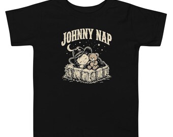 May include: A black short-sleeve t-shirt featuring a graphic print of a child in a cowboy hat sleeping on a hay bale with a teddy bear. Above the illustration, the text JOHNNY NAP is printed in a light beige, arched, serif font.