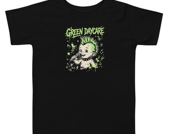 May include: A black short-sleeve t-shirt featuring a graphic print of a baby with a green mohawk holding a microphone. The text GREEN DAYCARE is printed in green above the baby, surrounded by green paint splatters and small icons.
