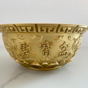 May include: A gold-colored bowl featuring a wide rim with a geometric Greek key pattern. The exterior is embossed with Chinese characters, stylized ingot shapes, and floral motifs. The base of the bowl has a petal-like design around the bottom edge.