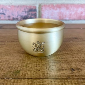 May include: A small, round, brushed gold-tone metal bowl with a smooth, curved rim. The exterior features an engraved character design in the center. The bowl has a matte finish and a consistent metallic gold color throughout its exterior surface.