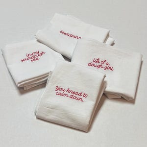May include: Four folded white kitchen towels featuring red embroidered text. The phrases include "in my sourdough era," "breadwinner," "life of a dough girl," and "you knead to calm down." Each towel is made of white fabric with red stitching.