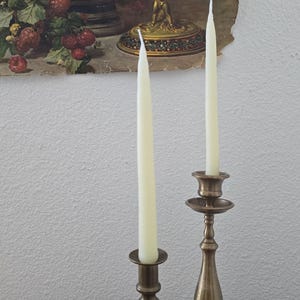 May include: Two tall, slender, off-white taper candles are held upright in separate brass candlesticks. The candlesticks feature a metallic finish with a tiered, rounded base and a flared top rim, standing on a wooden surface.
