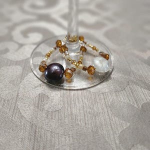 May include: A circular wine glass charm featuring a mix of amber-colored faceted beads, clear beads, a single dark purple round bead, and a single white round bead. The charm is constructed with a thin metal wire loop designed to fit around a stem.