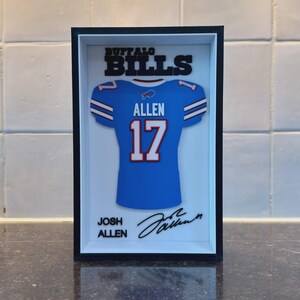 May include: A rectangular black shadow box frame containing a layered paper art piece of a blue Buffalo Bills jersey with the name ALLEN and number 17 in white. The text BUFFALO BILLS appears at the top, with JOSH ALLEN and a signature below.