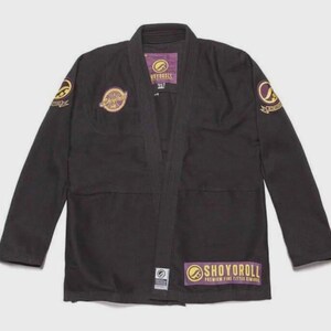 May include: A black martial arts gi jacket featuring long sleeves and a thick lapel. The garment displays yellow and purple circular patches on the chest and shoulders, with a rectangular patch at the bottom reading SHOYOROLL.