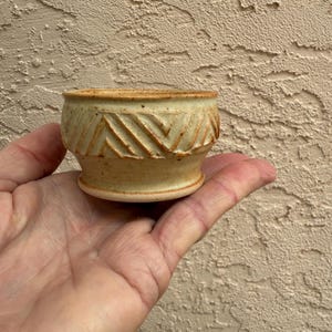 May include: A small, handmade ceramic bowl with a light beige glaze. The bowl features a decorative band of carved chevron patterns around the middle. The rim and base have a slightly darker, reddish-brown hue. The bowl is held in a hand.