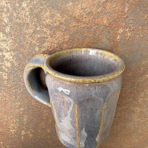 May include: A handmade ceramic mug with a unique, faceted design. The mug features a gray-purple glaze with yellow accents on the rim and vertical lines. The handle is a matching gray-purple color. A rustic, handcrafted piece.