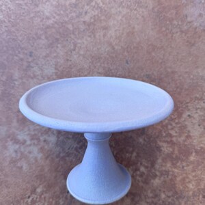 May include: A small, round, light purple cake stand with a pedestal base. The stand has a textured surface and is made of ceramic or a similar material. The stand is approximately 6 inches in diameter and 4 inches tall.