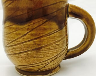 May include: A ceramic mug with a glossy, honey-brown glaze featuring a textured surface of diagonal, incised lines. The mug has a rounded body, a sturdy curved handle, and a slightly flared rim, showcasing a warm, earthy color palette.