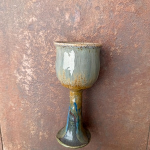 May include: A handmade ceramic goblet with a wide bowl and a slender stem. The goblet features a blend of colors, including blue, gray, and brown, with a textured surface. The base is dark gray, and the stem has blue and yellow accents.