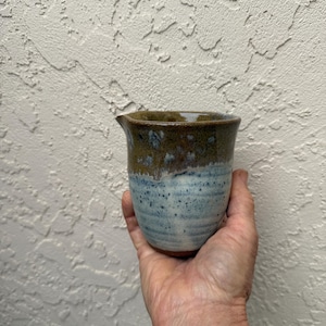 May include: A handmade ceramic creamer with a spout, held in a hand. The creamer has a speckled blue and brown glaze. The top is brown, transitioning to blue with white speckles. The creamer is a unique piece of pottery.