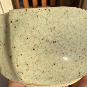 May include: A light green ceramic bowl with a textured, crackled surface and small brown speckles. The bowl has a unique, slightly irregular shape, suggesting a handmade quality. The interior is smooth and glossy.
