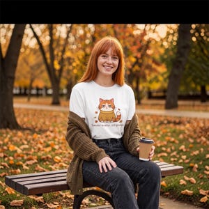 May include: A white t-shirt featuring a graphic of an orange tabby cat wearing a yellow sweater, surrounded by falling autumn leaves. Below the cat, the text reads, "Sweater weather, still grumpy." The shirt is paired with dark jeans and a brown cardigan.