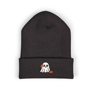 May include: A charcoal grey knit beanie featuring an embroidered graphic on the folded cuff. The embroidery depicts a small dog wearing a white ghost costume, with orange ears visible, standing next to a small orange pumpkin on the dark fabric.