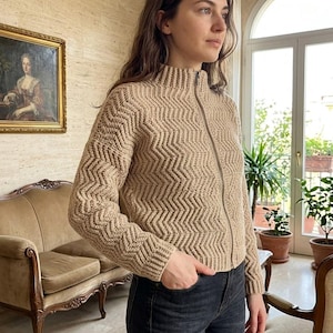 Peut inclure: Un gilet zippé en tricot beige avec un motif chevron texturé, un col montant et des manches longues. Le vêtement est porté avec un jean en denim gris foncé. Le gilet présente une fermeture éclair intégrale et des poignets côtelés.