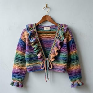 Puede incluir: Un cárdigan de ganchillo corto de manga larga con un degradado de arcoíris vibrante. La prenda incluye un cuello con volantes y detalles bordados en forma de hoja, un cierre frontal con lazos y puños con volantes sobre una percha de madera.