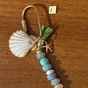 May include: A gold-toned metal keychain featuring a white seashell, a small gold-toned starfish charm, a green tassel, and a vertical strand of blue, white, and iridescent beads. A small yellow square paper with the handwritten number 1 is visible.