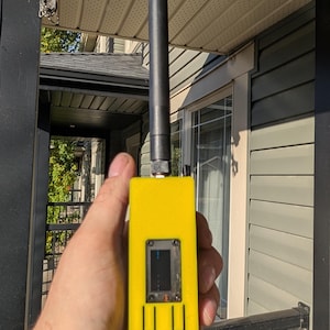 Op de afbeelding: Een felgeel rechthoekig elektronisch apparaat met een zwarte antenne aan de bovenkant, een klein rechthoekig scherm in het midden en twee verticale rechthoekige uitsparingen onder het scherm aan de voorzijde.