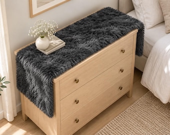 May include: A dark gray faux fur runner draped over a light wood dresser with three drawers. The runner has a long, shaggy texture and hangs over the edges of the dresser. The dresser features small, round, metallic knobs on each drawer.