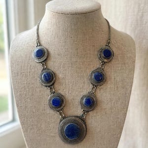 May include: A silver-toned necklace featuring seven circular medallions with intricate metalwork patterns and deep blue circular stones. The largest medallion is centered at the bottom, with three smaller medallions ascending on each side of the chain.