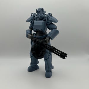May include: A gray plastic action figure of a soldier in full tactical power armor, featuring a helmet with a circular breathing apparatus and shoulder pauldrons. The figure holds a black multi-barreled rotary machine gun in its hands.