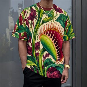 May include: A short-sleeve t-shirt featuring a vibrant, all-over botanical print of green and deep red Venus flytrap plants against a cream-colored background. The shirt has a solid dark red crew neckline and is paired with plain black trousers.