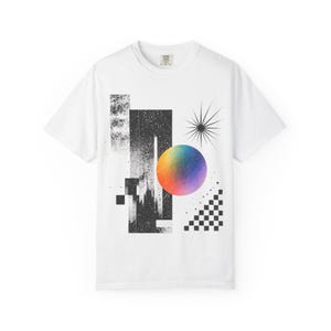 May include: A white short-sleeve t-shirt featuring a graphic design with a central gradient sphere transitioning from orange to purple. The print includes black rectangular shapes with distressed textures, a starburst icon, and a checkered pattern.