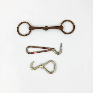 May include: A collection of three metal tools: a brown rusted horse bit with two circular rings, a red and silver metal hook tool with a looped handle, and a small silver-toned metal hook with a hinged loop mechanism.