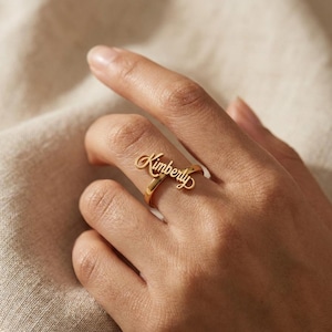 Könnte beinhalten: Ein goldfarbener Metallring mit dem Namen Kimberly in einer kursiven Schriftart. Der Name ist horizontal über der Oberseite des Rings positioniert, der aus einem einfachen, glatten und polierten goldfarbenen Metall besteht.