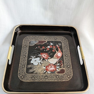 May include: A rectangular black serving tray with a gold-toned rim and light-colored wrapped handles. The center features a detailed illustration of a bird, red and white flowers, and foliage, all enclosed within an intricate gold-colored border.