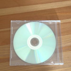 May include: A clear plastic CD jewel case containing a standard circular compact disc. The disc has a reflective, iridescent silver surface with a white center hub. The case is square with a transparent cover, revealing the disc inside clearly.