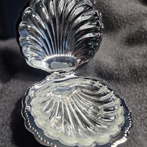 May include: A silver-toned metal trinket box shaped like an open seashell. The interior and exterior feature a ribbed, scalloped design with a polished, reflective finish. The two halves are connected by a small metal hinge at the base.