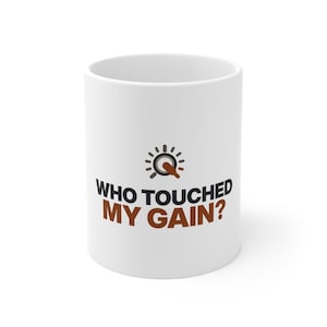 May include: A white ceramic mug featuring a graphic of a circular audio gain knob icon above the black and brown text that reads WHO TOUCHED MY GAIN? The design is centered on the front of the mug, which has a smooth, cylindrical body.
