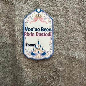 Peut inclure: Une &eacute;tiquette cadeau rectangulaire &agrave; bord festonn&eacute; orn&eacute;e d'une couronne dor&eacute;e, d'un ruban rose et d'un ch&acirc;teau bleu. Le texte indique You've Been Pixie Dusted! et From: en lettres bleu fonc&eacute; et roses sur fond cr&egrave;me.