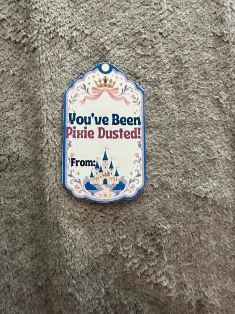 Peut inclure: Une &eacute;tiquette cadeau rectangulaire &agrave; bord festonn&eacute; orn&eacute;e d'une couronne dor&eacute;e, d'un ruban rose et d'un ch&acirc;teau bleu. Le texte indique You've Been Pixie Dusted! et From: en lettres bleu fonc&eacute; et roses sur fond cr&egrave;me.