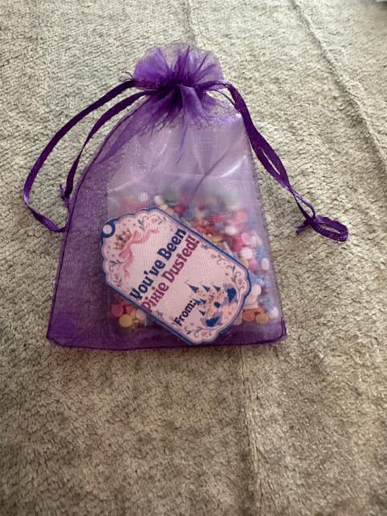Peut inclure: Un sachet en organza violet avec cordon de serrage, rempli de petites perles circulaires multicolores. Une &eacute;tiquette d&eacute;corative est fix&eacute;e &agrave; l'avant, avec une illustration de ch&acirc;teau et le texte : You've Been Pixie Dusted! From: