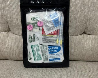 Peut inclure: Une pochette refermable noire contenant diverses fournitures de premiers secours, notamment des compresses Curad, de la crème à l'hydrocortisone, des lingettes Boo Boo, deux cotons-tiges à embouts jaunes et deux petits comprimés roses ronds avec du texte.
