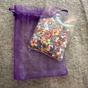 Peut inclure: Un sachet en plastique transparent rempli d'un assortiment color&eacute; de petites d&eacute;corations sucr&eacute;es rondes et en forme d'&eacute;toile dans des tons rose, jaune, bleu et blanc, pos&eacute; sur un sac cadeau rectangulaire en organza violet.