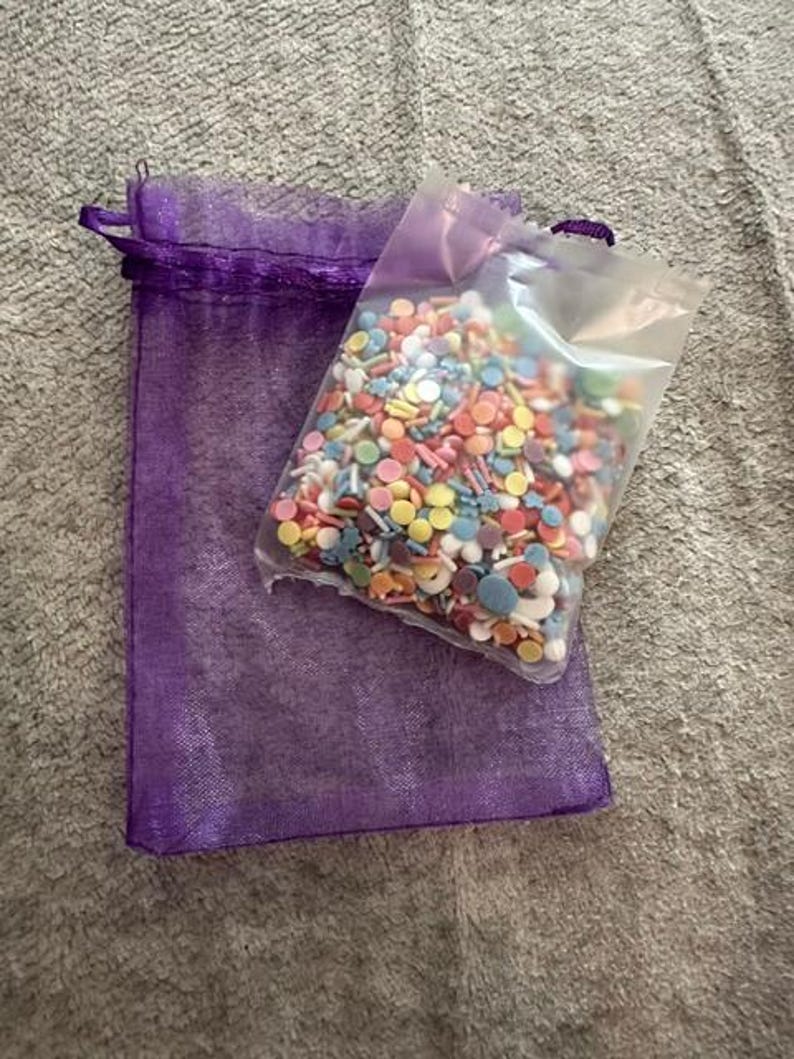 Peut inclure: Un sachet en plastique transparent rempli d'un assortiment color&eacute; de petites d&eacute;corations sucr&eacute;es rondes et en forme d'&eacute;toile dans des tons rose, jaune, bleu et blanc, pos&eacute; sur un sac cadeau rectangulaire en organza violet.