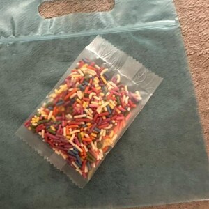 Peut inclure: Un petit sachet en plastique transparent rempli de vermicelles en sucre multicolores dans des tons de rouge, orange, jaune, vert, bleu et violet, pos&eacute; sur un grand sac en plastique bleu sarcelle translucide avec une poign&eacute;e d&eacute;coup&eacute;e.