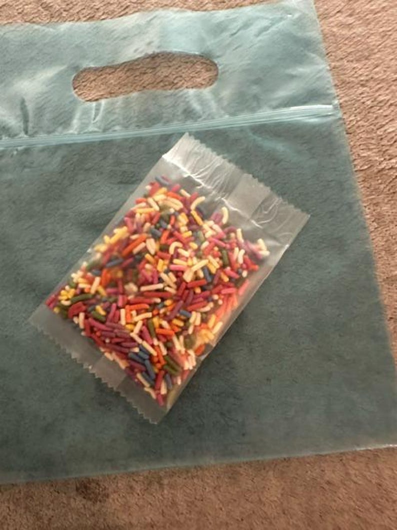 Peut inclure: Un petit sachet en plastique transparent rempli de vermicelles en sucre multicolores dans des tons de rouge, orange, jaune, vert, bleu et violet, pos&eacute; sur un grand sac en plastique bleu sarcelle translucide avec une poign&eacute;e d&eacute;coup&eacute;e.