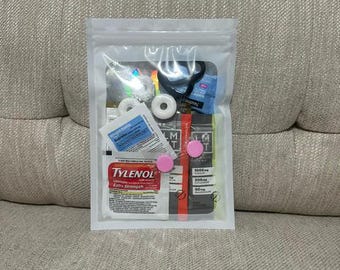 Peut inclure: Une pochette refermable en plastique transparent contenant divers petits articles, dont un sachet de Tylenol Extra Strength, un sachet d'ibuprofène Amazon Basics, deux pastilles rondes blanches, un élastique noir et un sachet LMNT avec électrolytes.