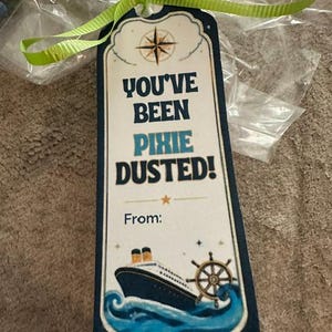 Peut inclure: Une &eacute;tiquette cadeau rectangulaire avec une bordure bleu fonc&eacute;, attach&eacute;e par un ruban vert lime. Elle pr&eacute;sente une rose des vents, un paquebot sur des vagues bleues, une barre de navire et le texte : YOU'VE BEEN PIXIE DUSTED! From: en bleu.