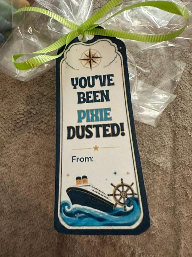 Peut inclure: Une &eacute;tiquette cadeau rectangulaire avec une bordure bleu fonc&eacute;, attach&eacute;e par un ruban vert lime. Elle pr&eacute;sente une rose des vents, un paquebot sur des vagues bleues, une barre de navire et le texte : YOU'VE BEEN PIXIE DUSTED! From: en bleu.