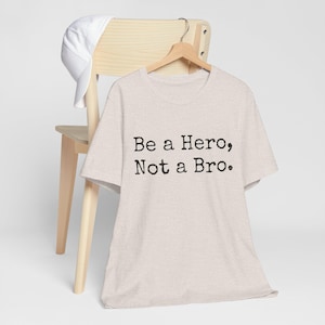 May include: A heathered beige short-sleeve t-shirt featuring the black text "Be a Hero, Not a Bro." printed on the front. The shirt is displayed on a wooden hanger, and a plain white baseball cap is draped over the back of the wooden chair.